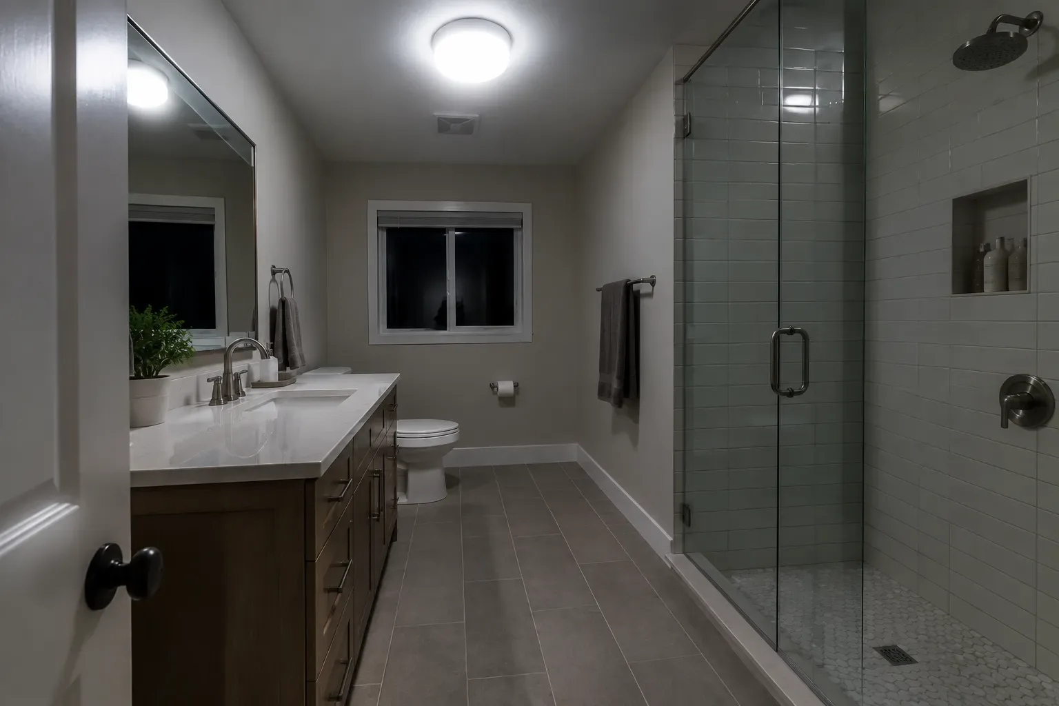 Eye-level wide view of an existing bathroom with a vanity and mirror on the left, toilet beneath a window at the back, a glass-enclosed tiled shower on the right, and a bright central ceiling light illuminating the room.